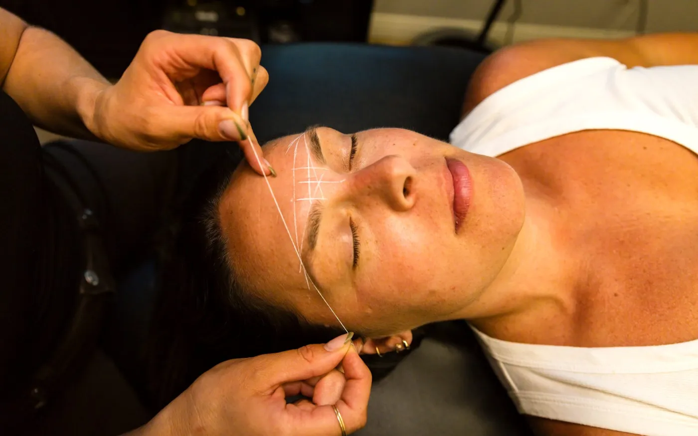 An artist maps a client's brows with white pencil and a mapping thread.
